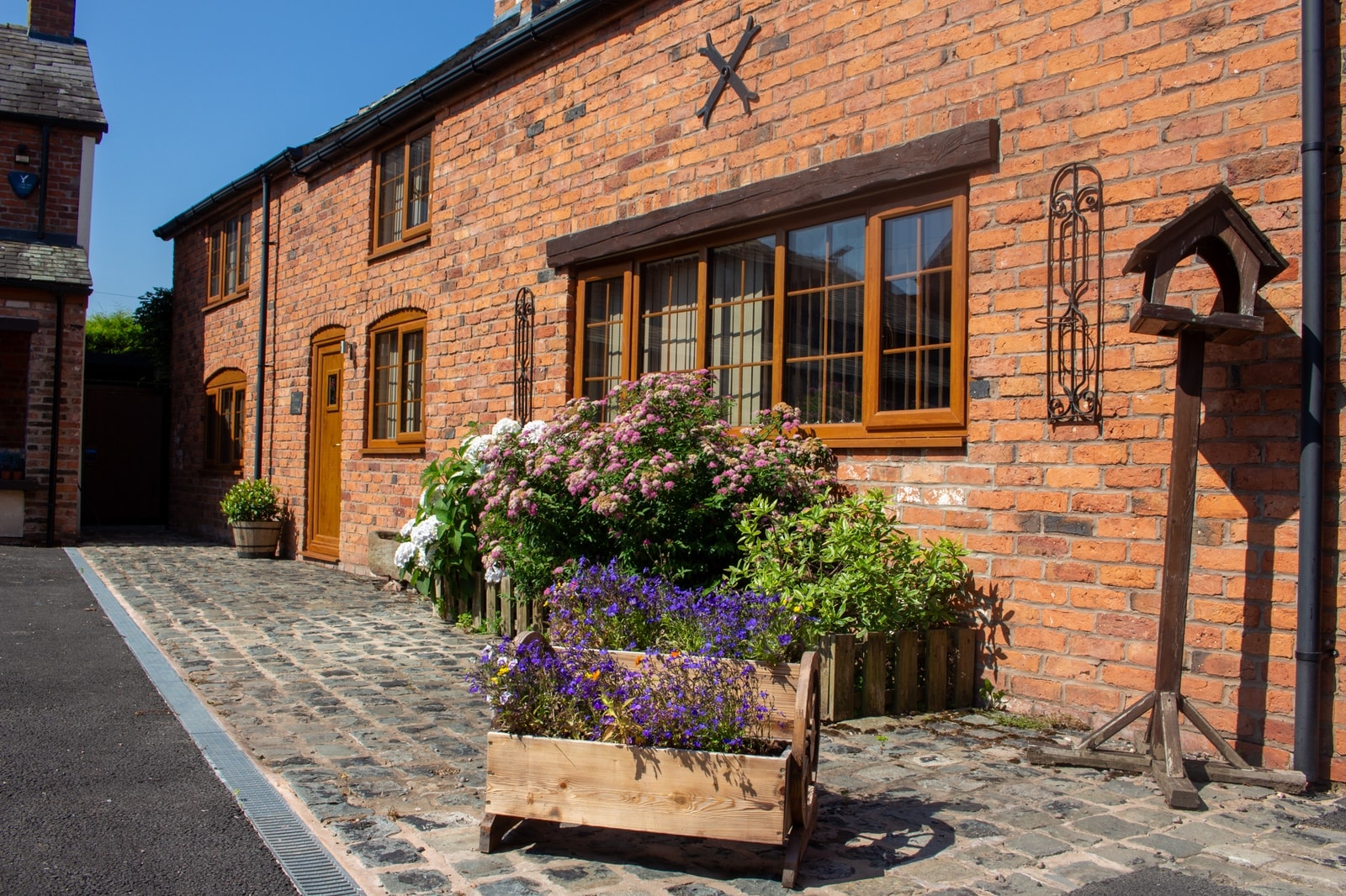 A cottage frontage in warm brick, planters and a hanging basket in full flower beside the oak door