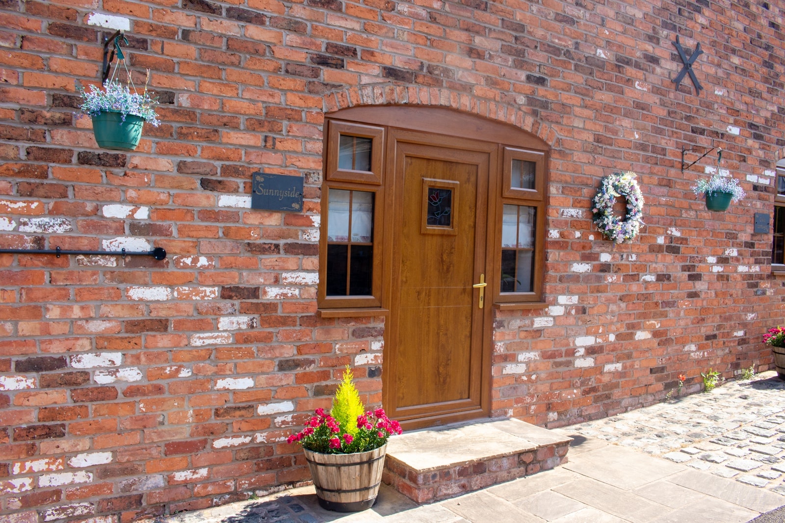 An oak door framed by hanging baskets, a wreath and a planter on the step